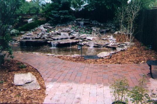 Brick patio leads to a pond with a waterfall, surrounded by rocks and mulch; trees in background.