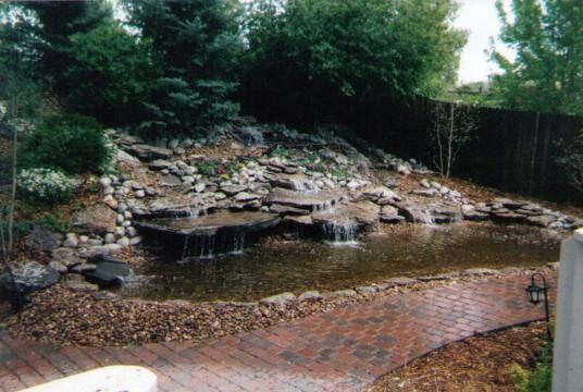 Water flows over rocks into a pond, surrounded by greenery and a brick pathway.