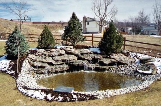A backyard pond with a small waterfall, rocky edges, and evergreens, partially covered in snow.