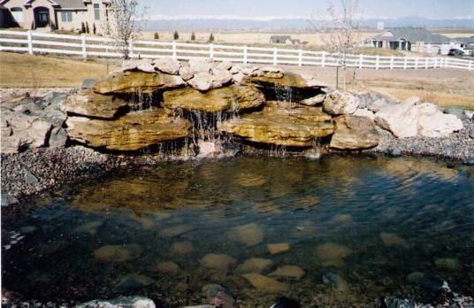 Water fountain with waterfall cascading into a pond. Brown rocks surround it, with a white fence and houses in the background.