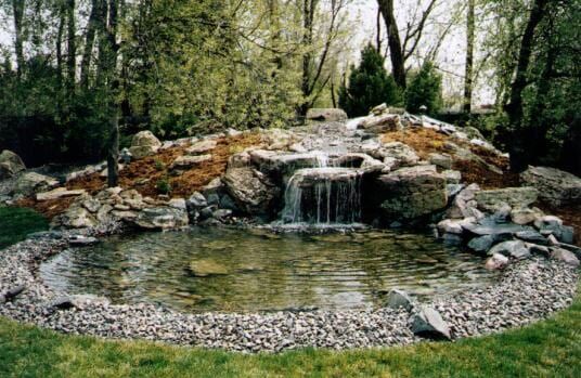 Pond with waterfall surrounded by rocks and greenery, in a backyard setting.