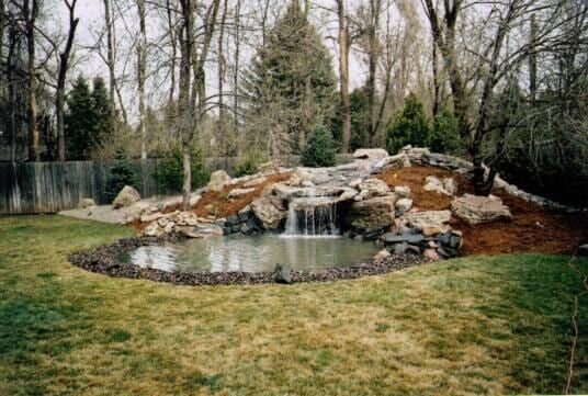 Backyard pond with waterfall cascading into a small body of water, surrounded by rocks and grass.