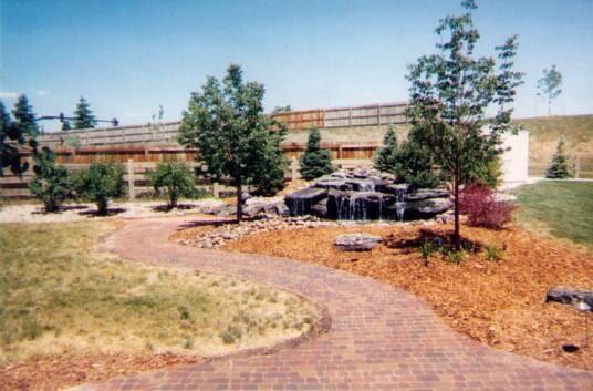 Brick pathway winds past a small waterfall feature and trees on a sunny day.