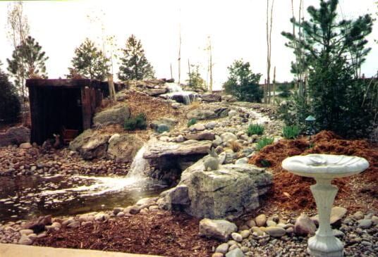 A small waterfall cascades over rocks in a garden with a bird bath and small building.