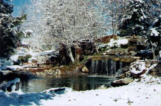 Snow-covered waterfall cascading into a pond, surrounded by snow-laden trees and rocks.