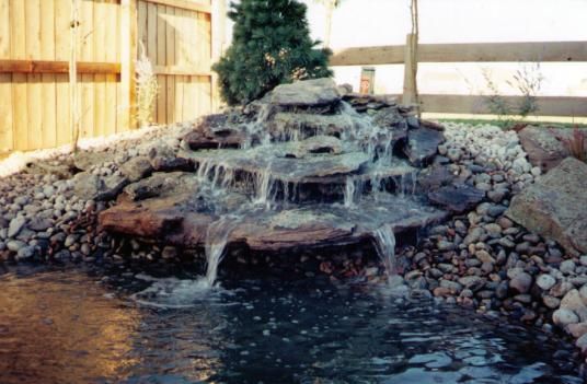 Water cascading down a tiered rock waterfall into a small pond, surrounded by rocks and a wooden fence.