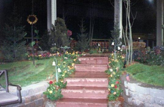 Brick stairs lined with flowers lead to a dark area with trees. A wreath is on the porch.