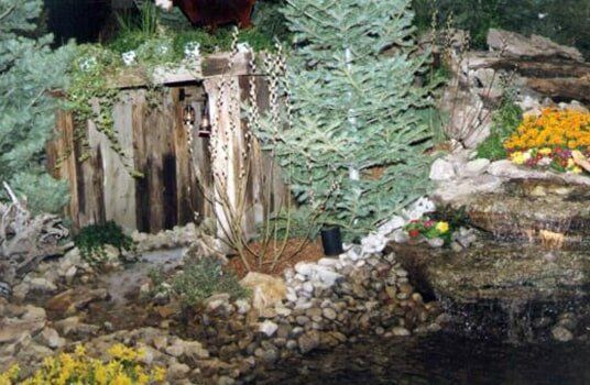 A decorative outdoor water feature with a wooden structure, waterfall, plants, and rocks.