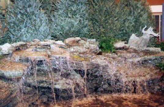 Waterfall with a light-colored sculpture on top; cascading water, rocks, and green foliage.