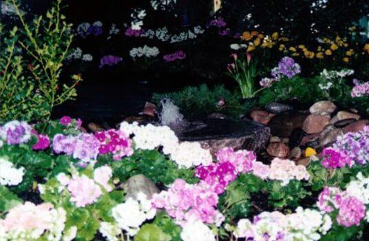 A vibrant garden with pink, purple, and white flowers surrounding a small fountain.