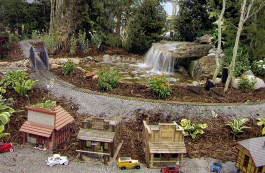 Miniature western town with waterfall, pond, and train tracks in a garden setting.