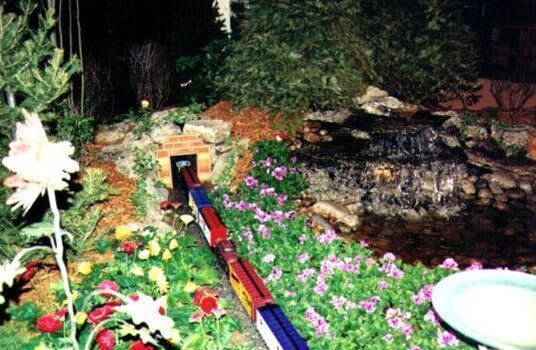 A toy train travels through a miniature landscape of flowers, a waterfall, and a tunnel.
