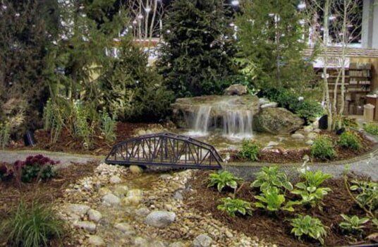 A miniature waterfall flows under a bridge, surrounded by plants and trees.