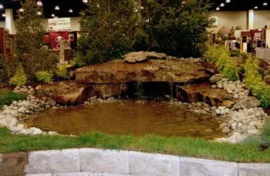 Small pond with a waterfall and landscaping, featuring rocks, trees, and grass.