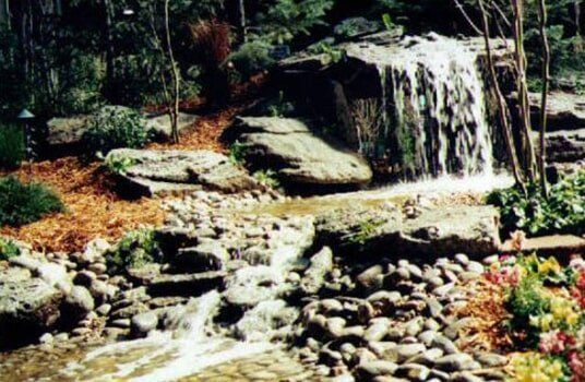 Waterfall cascading over rocks, surrounded by lush green plants and flowers in a garden setting.