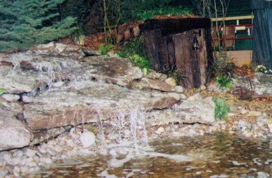 Waterfall cascading over rocks into a pond, with a wooden structure and greenery in the background.