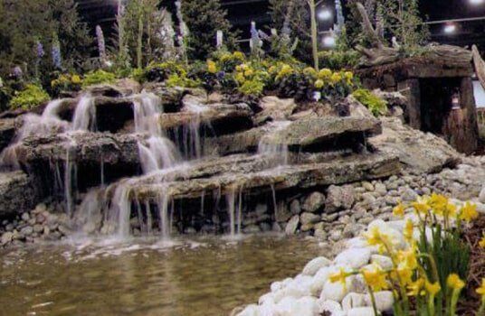 Waterfall cascading into a pond, surrounded by rocks, vegetation, and yellow flowers.