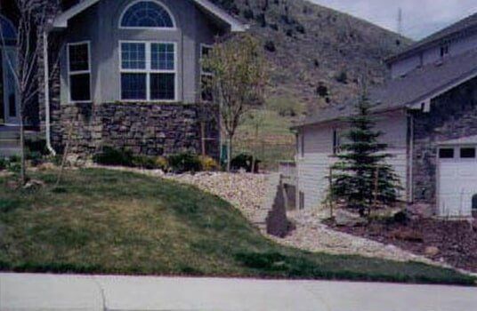 House with stone facade, arched window, and garage; sloped lawn with rock landscaping in front of mountain.