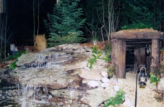 Waterfall cascading over rocks next to a wooden entrance with lanterns in a garden setting.