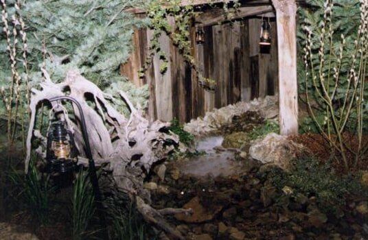 Rustic outdoor scene: weathered wooden structure, water feature, large white root, lanterns, greenery.