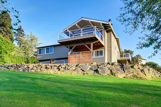 Modern hillside home with large deck, stone retaining wall, and green lawn under clear blue sky view.
