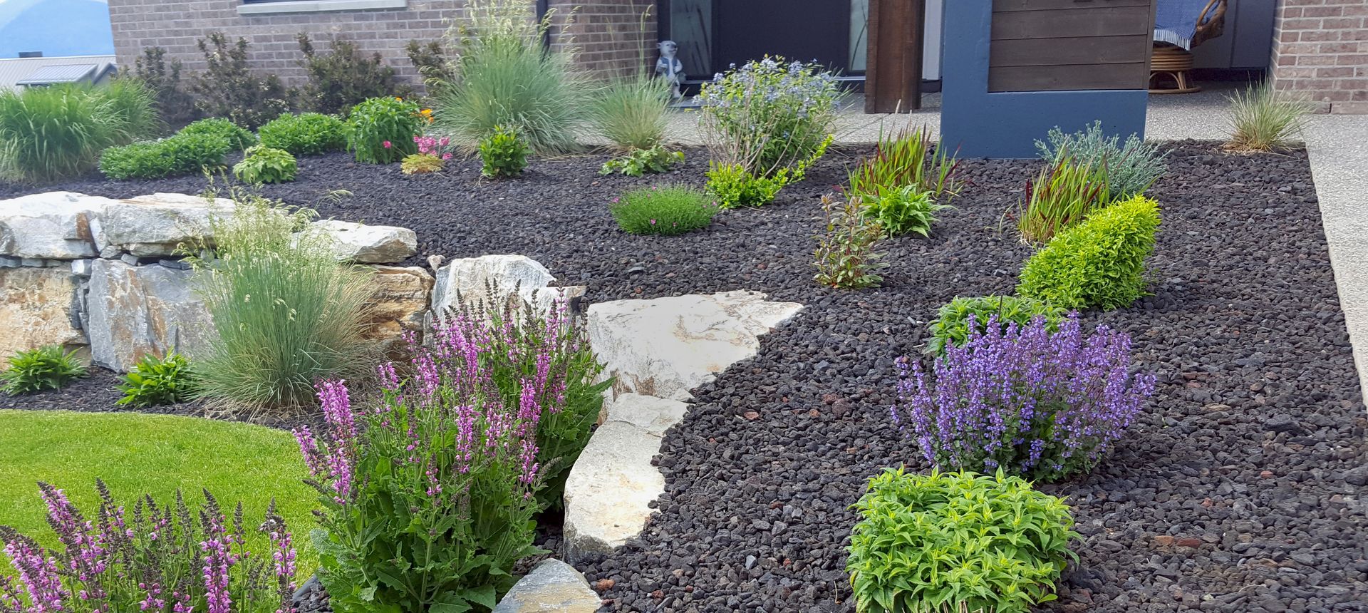 A garden bed with dark mulch, various green and purple plants, and stone accents in front of a house.