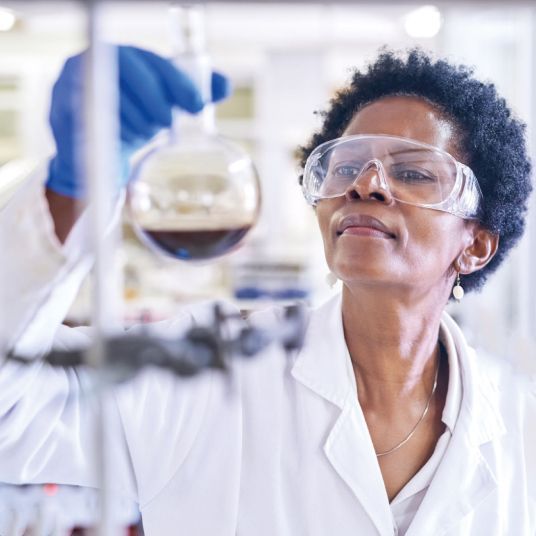 A woman in a lab coat and goggles is looking at a beaker