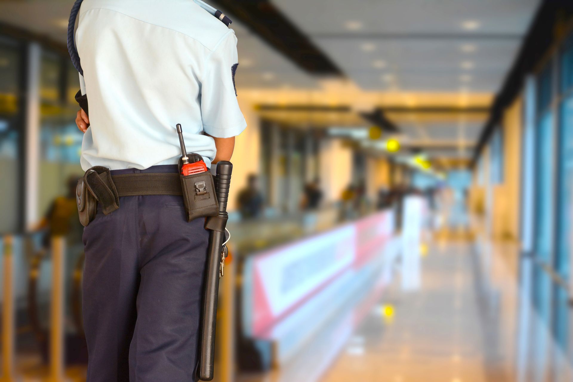 Security guard in uniform stands at an airport checkpoint, observing the terminal. He has a radio and baton on his belt.