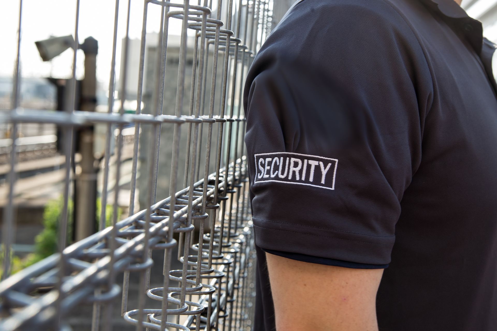 A man wearing a security shirt is standing next to a fence.