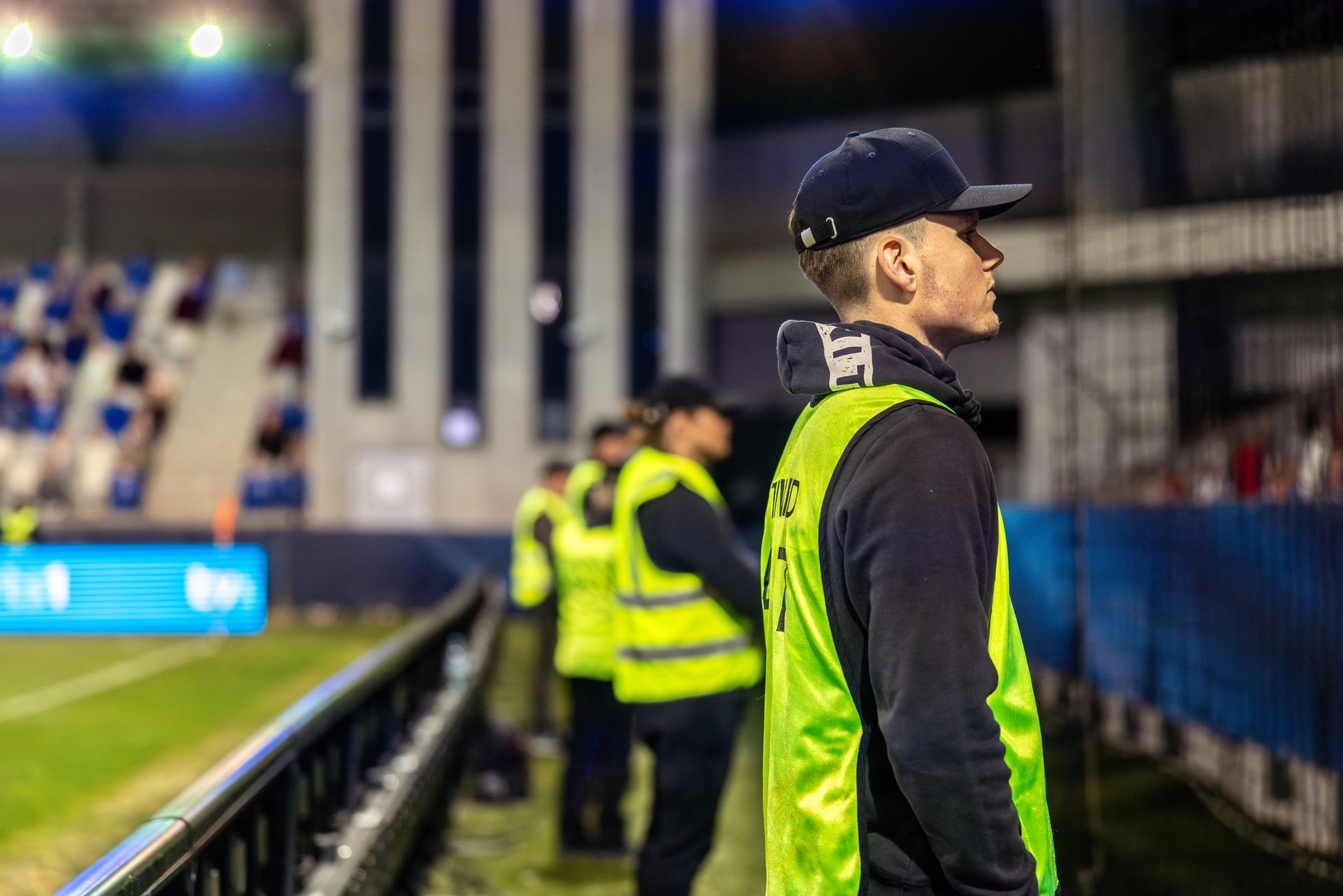 A man wearing a hat and a green vest is standing on a soccer field.