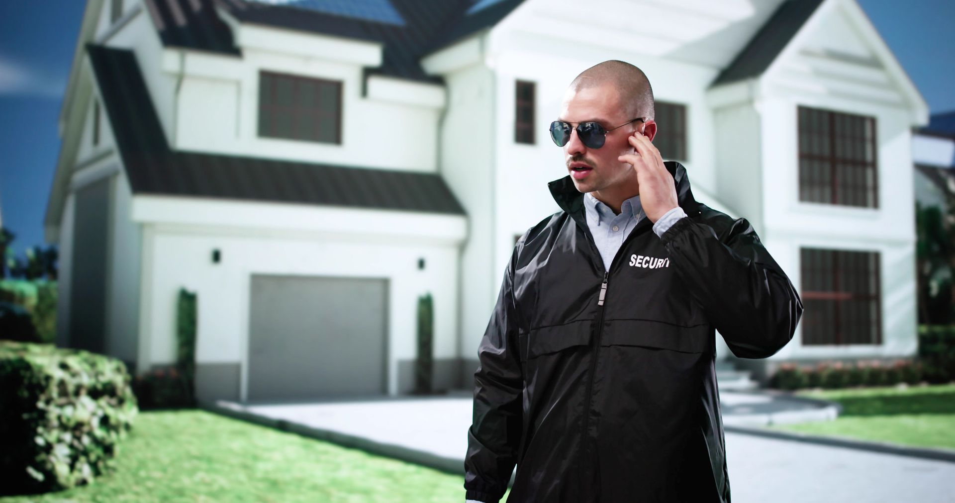 A man wearing sunglasses is talking on a cell phone in front of a house.