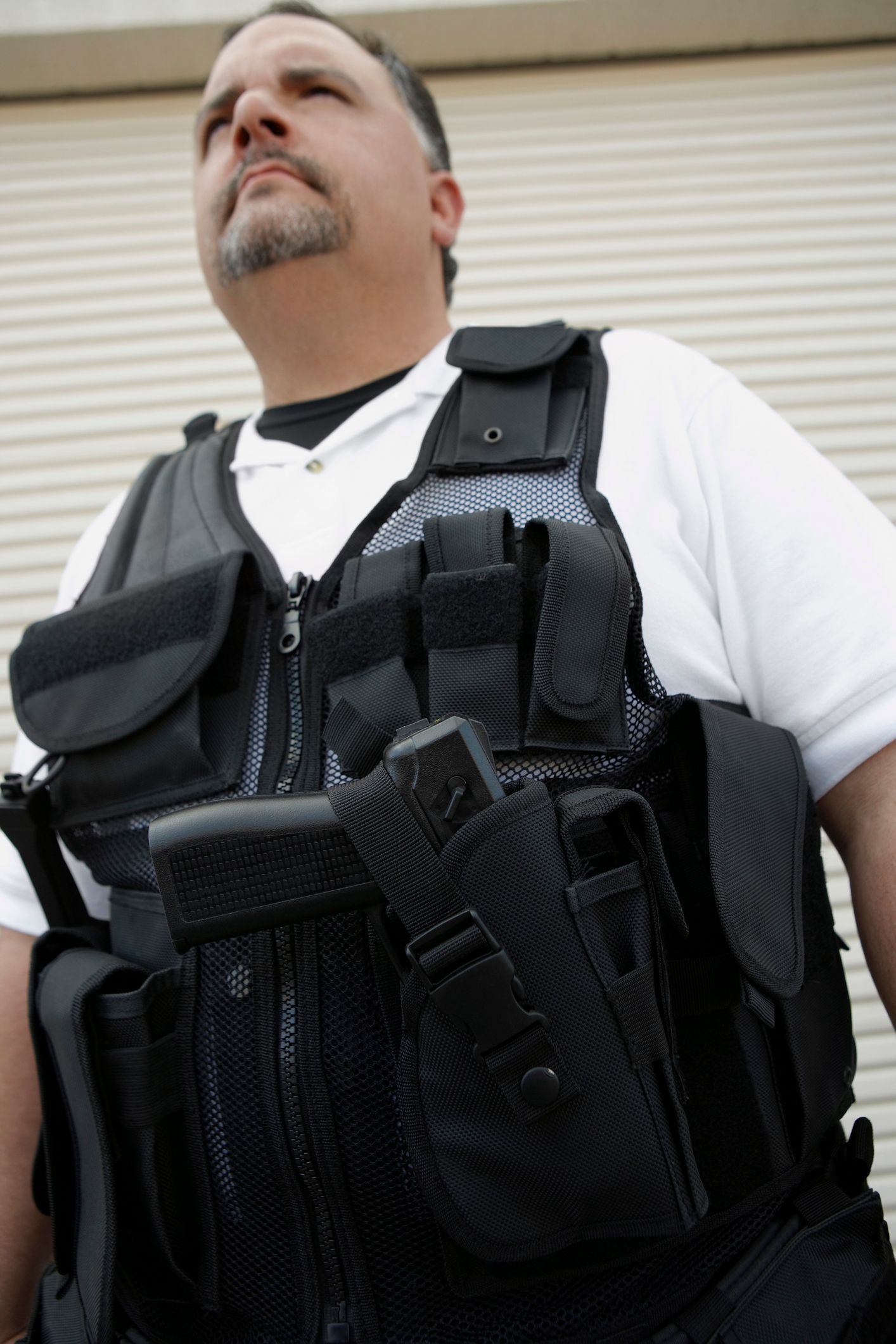 Man in a black tactical vest with a gun, looking up. Wearing a white shirt, standing outside a building.
