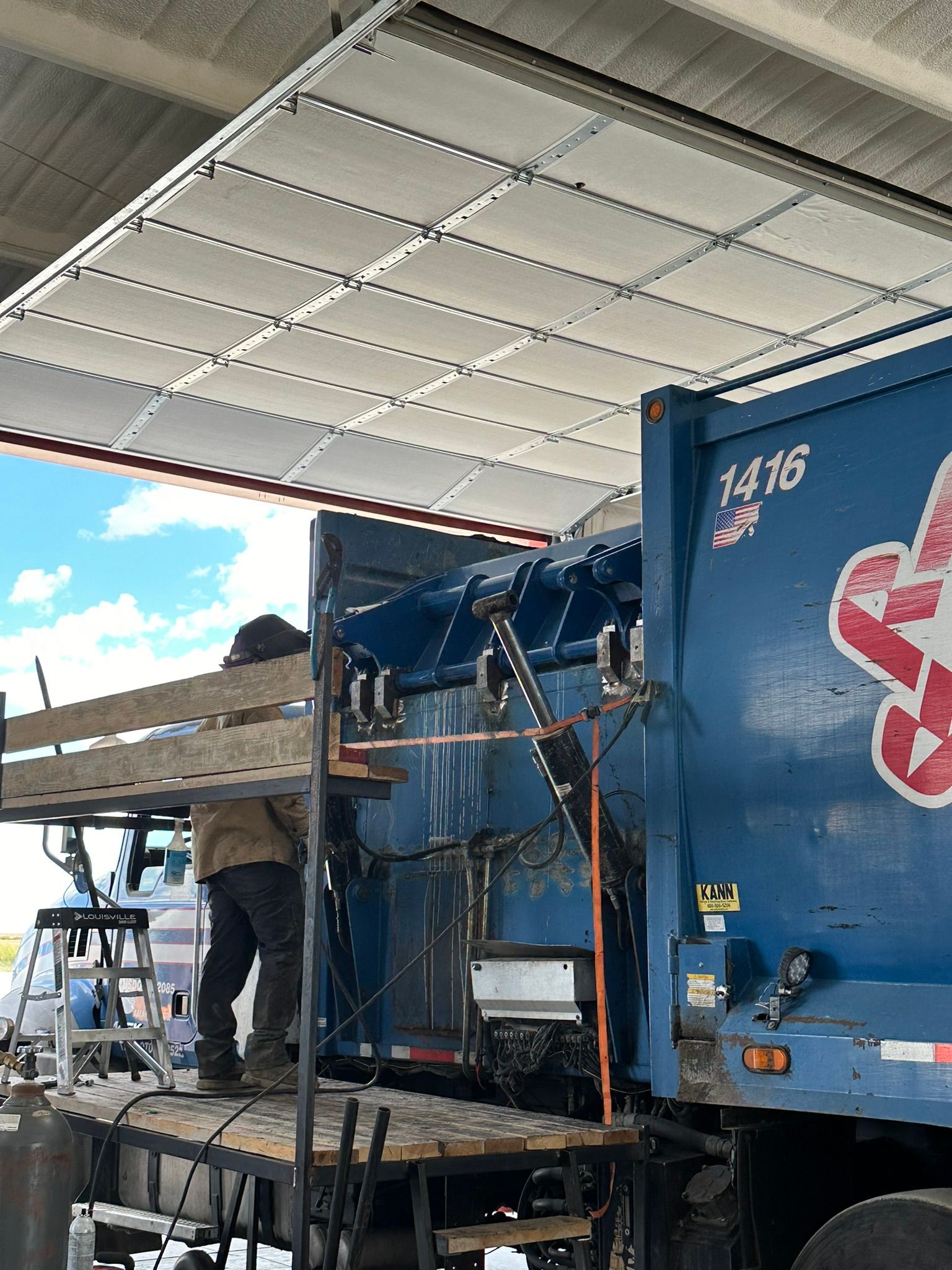 A blue garbage truck is being repaired in a garage.