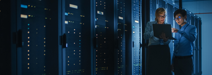 Two men are standing in a server room looking at a laptop.