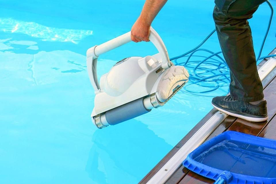 Person holding a white and gray robotic pool cleaner, lowering it into a blue pool.
