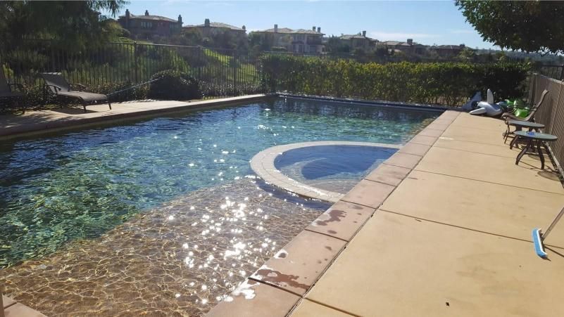 A pool with a built-in jacuzzi, concrete patio, and a view of houses in the background.