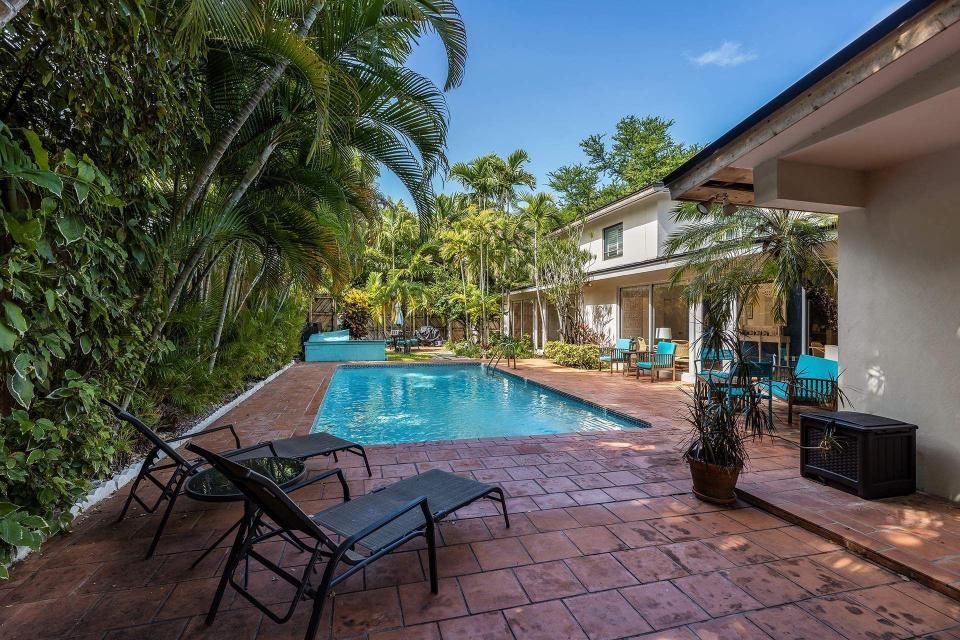 Pool and patio area of a house with lounge chairs, a table, and lush tropical greenery.