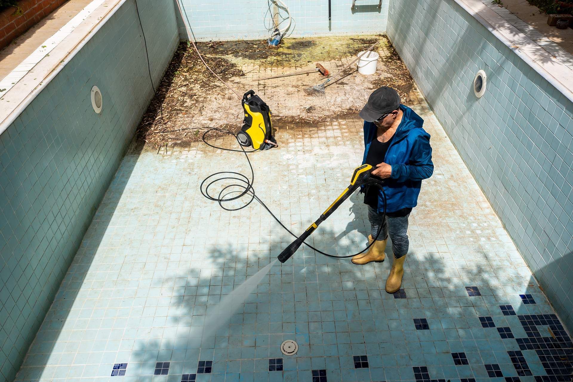 Two workers cleaning an empty empty pool, one spraying with a hose in a drained tiled basin.
