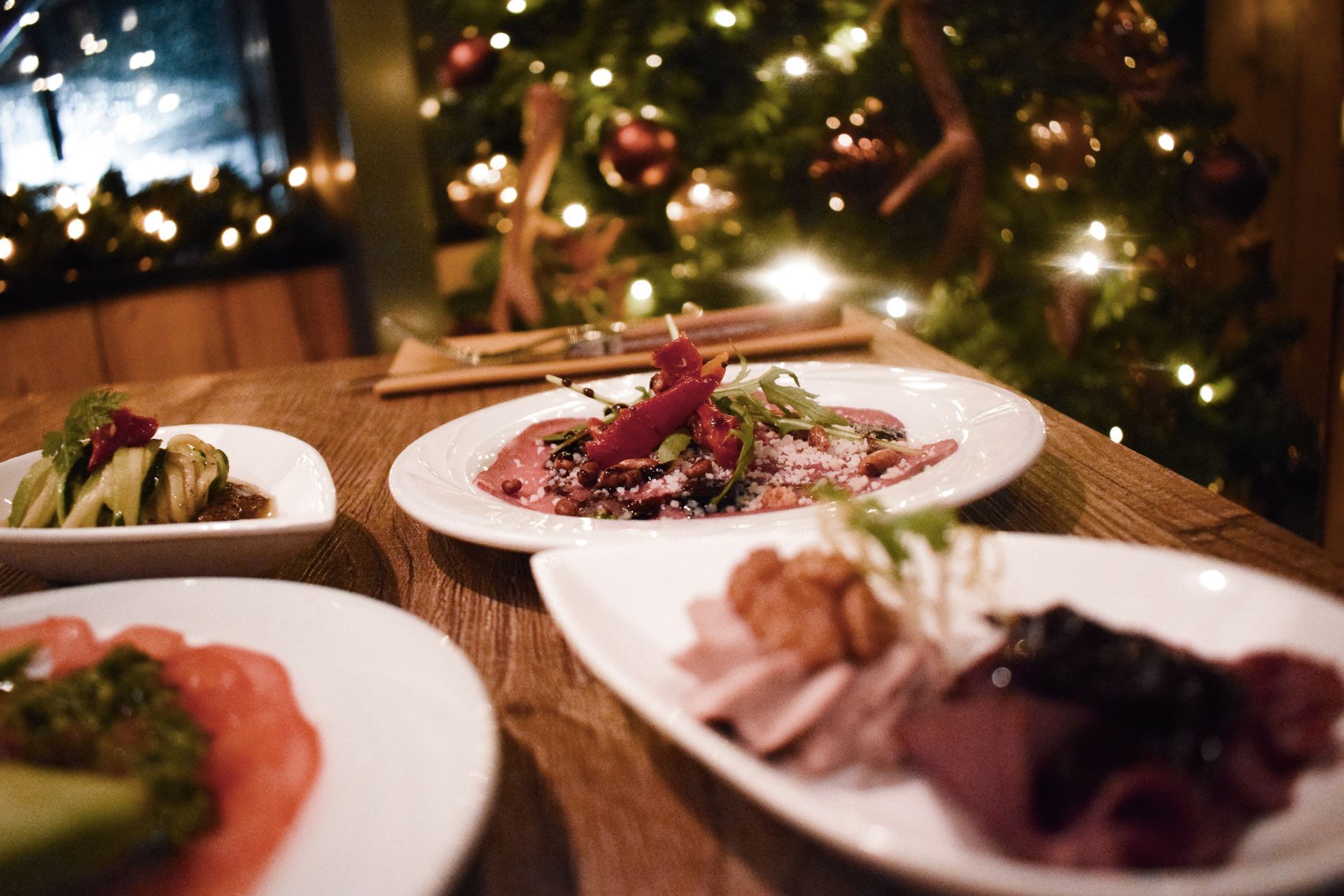 Borden met eten op een houten tafel, kerstboom met lichtjes op de achtergrond.