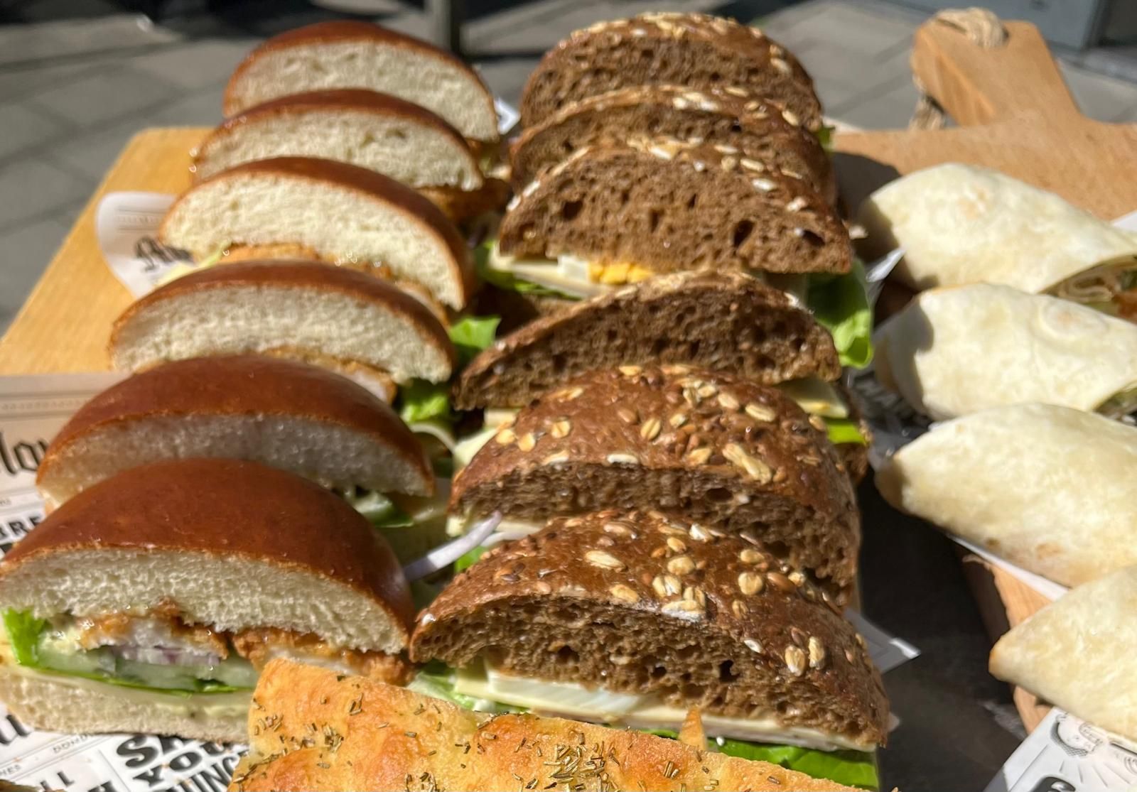 Sandwiches en wraps op een houten plank, zoals brioche- en roggebrood, met diverse vullingen.