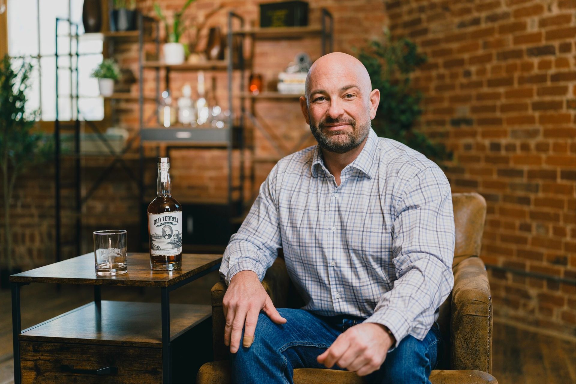 A person sitting in a leather chair near a side table with a bottle and glass, set against a rustic brick wall background.