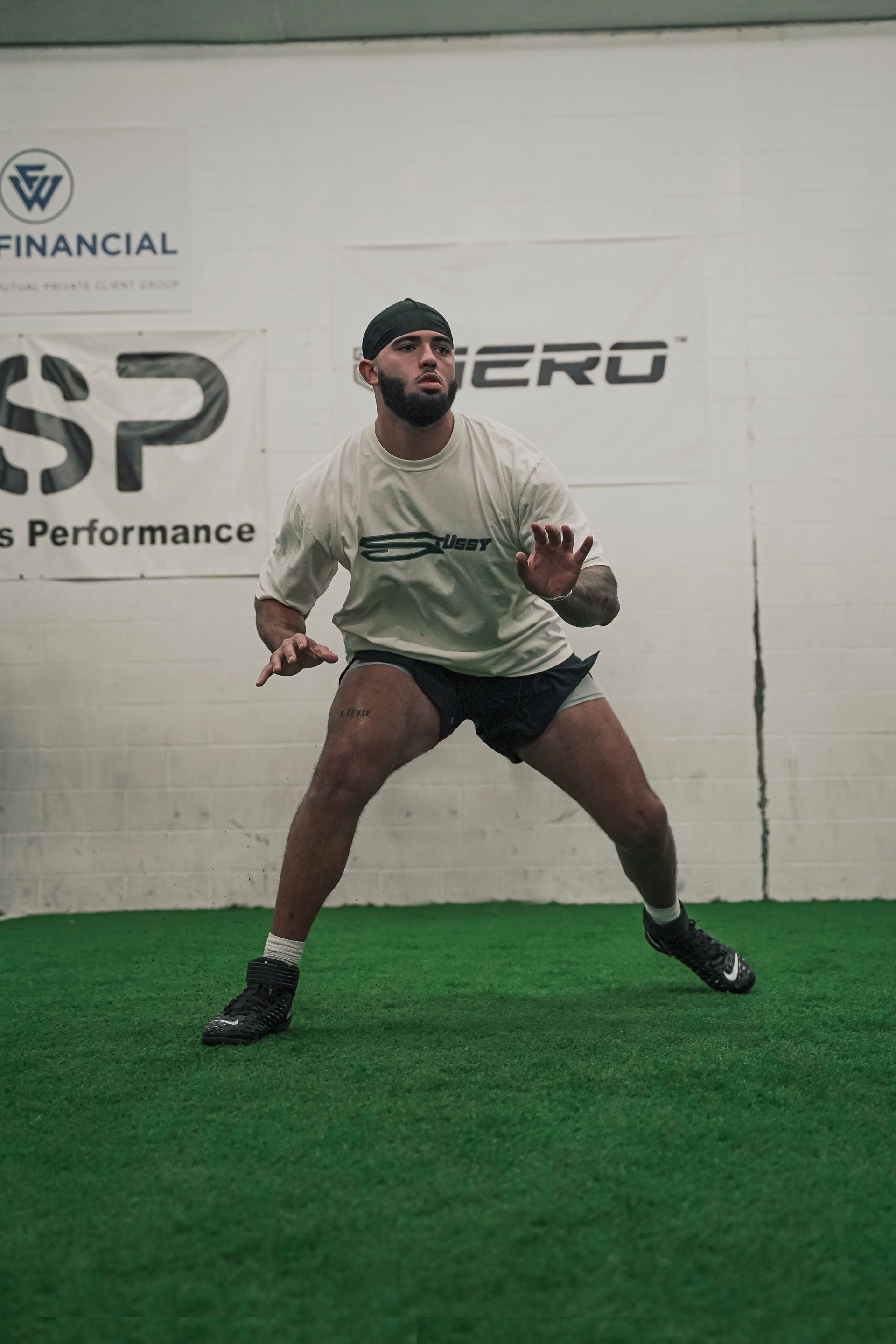 Athlete in athletic stance, moving laterally on artificial turf, wearing black shorts, t-shirt, and hat. Indoor gym setting.