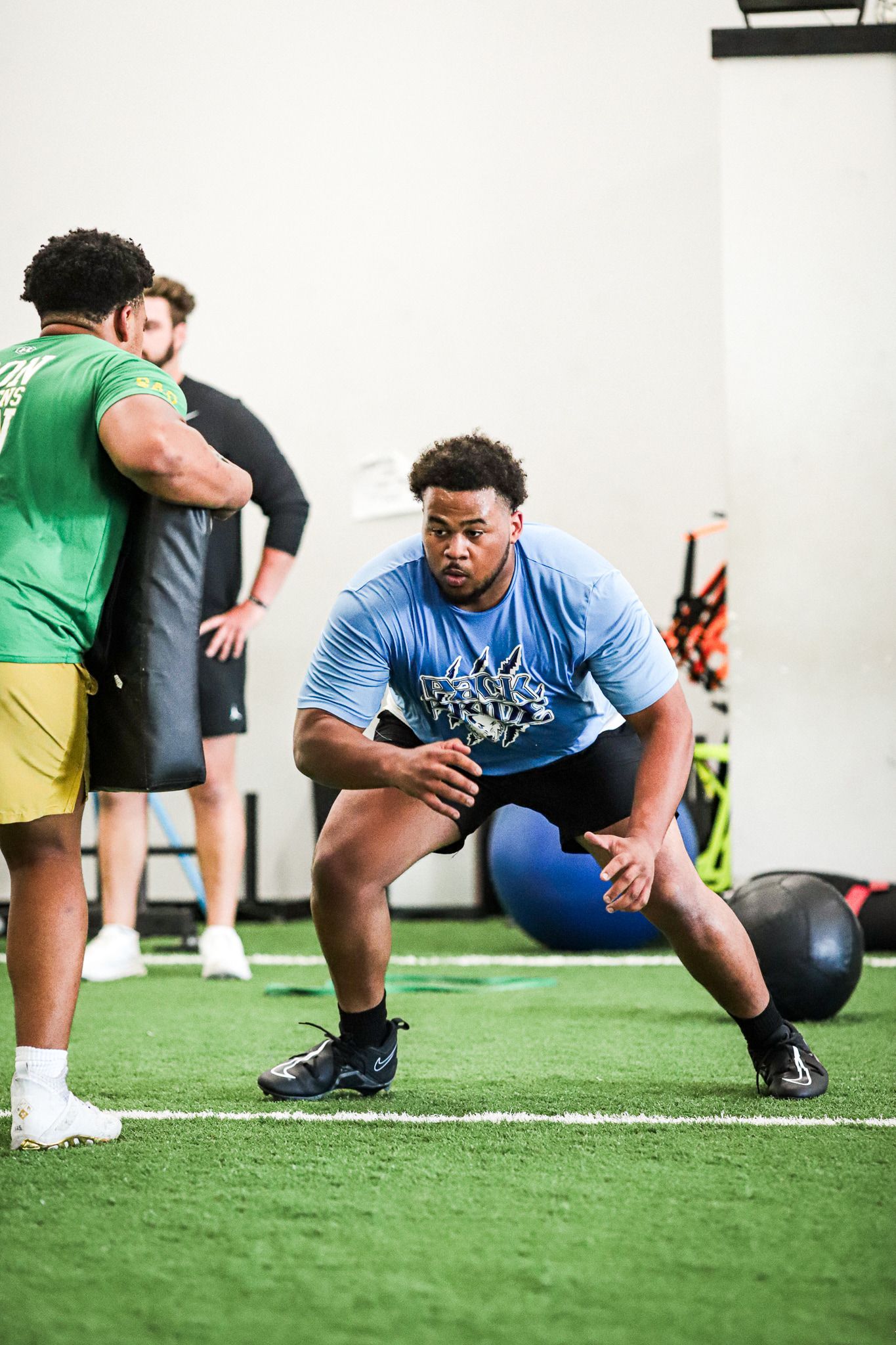 Football player in a blue shirt in a training stance, facing a trainer holding a pad. Gym setting.