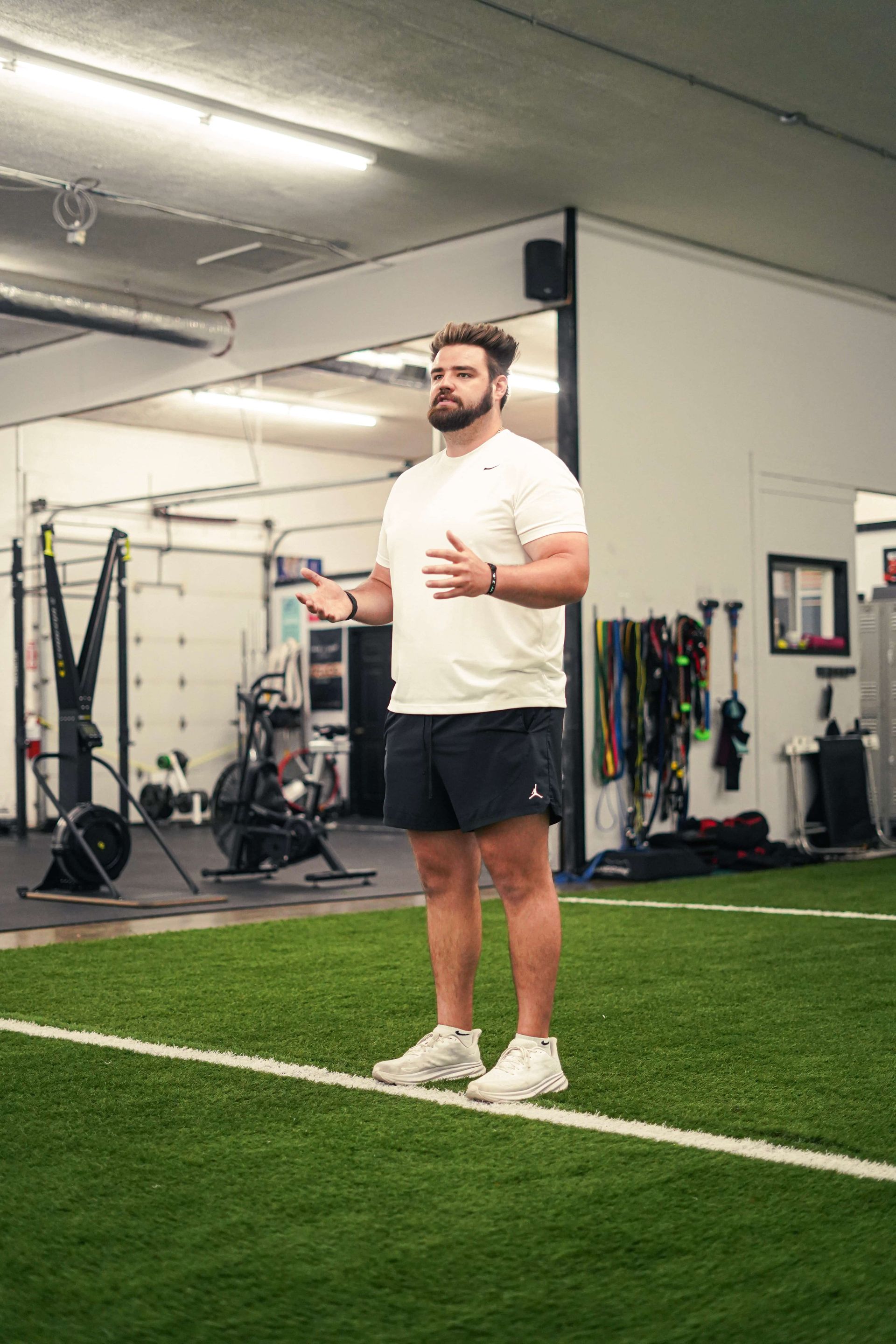 Man in gym, gesturing with hands, wearing white shirt, black shorts, and sneakers. Standing on turf.