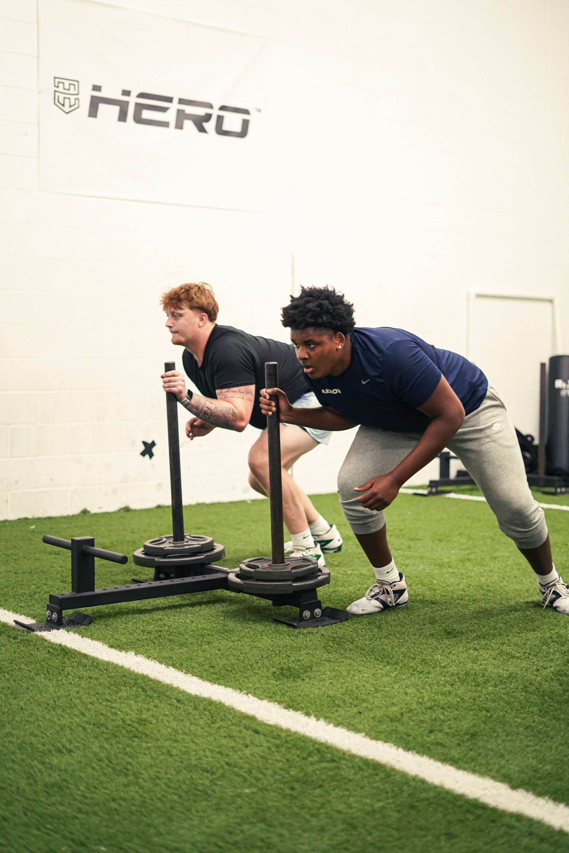 Two athletes push weighted sleds across artificial turf in a gym.