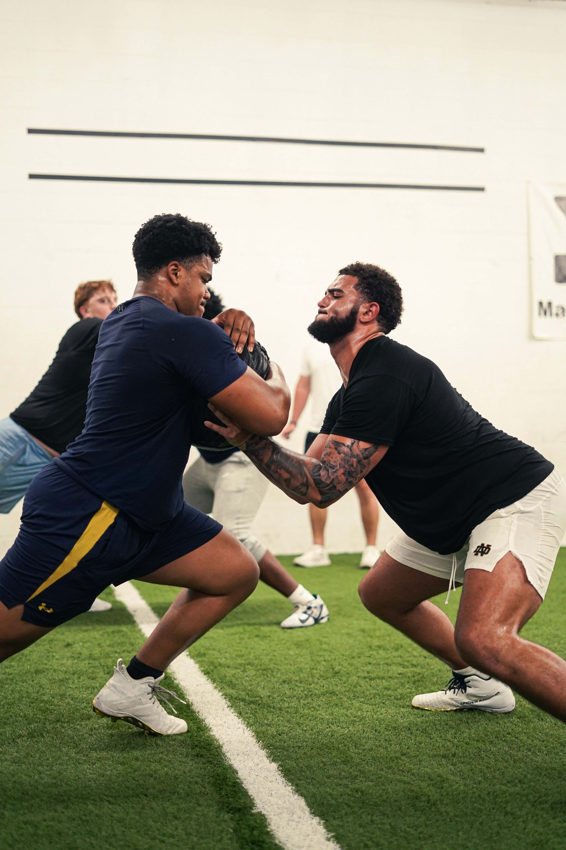 Two men face each other in athletic stance, practicing football drills in a gym.
