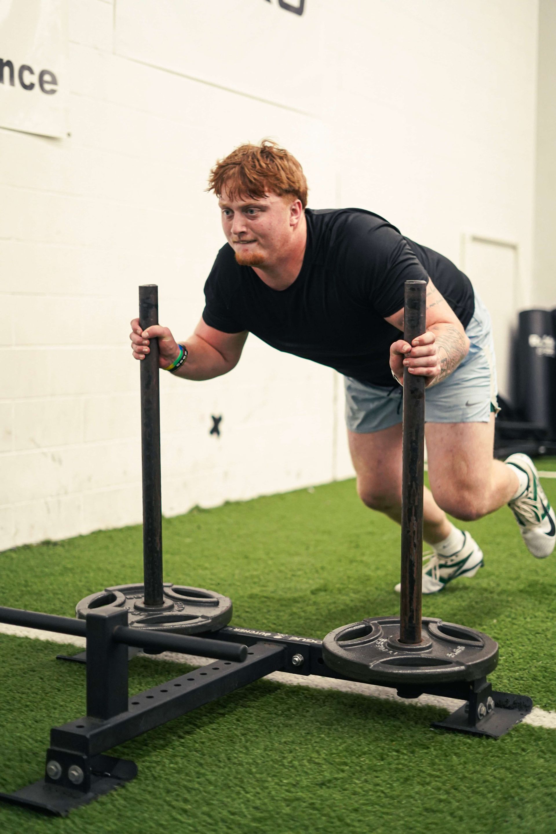Man in black shirt pushing a weighted sled on turf; indoors, gym setting.