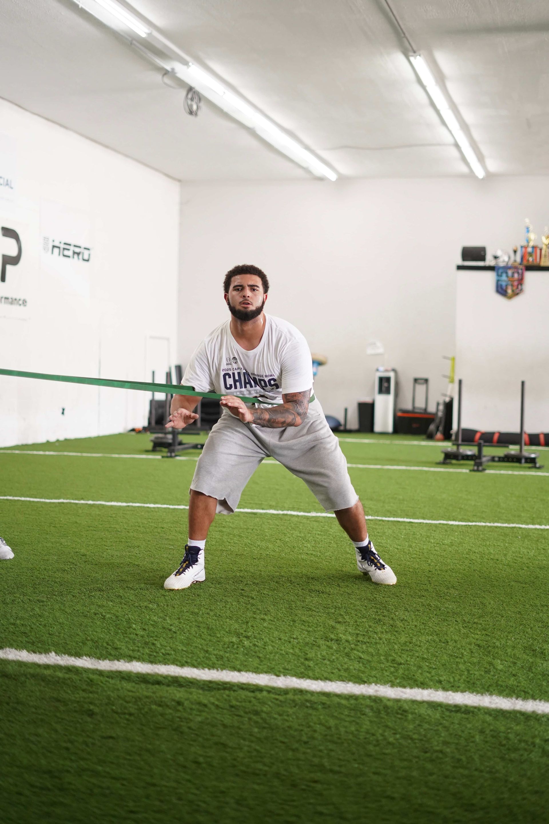 Man doing resistance band exercise on a turf field, holding band in front, wearing white t-shirt and shorts.
