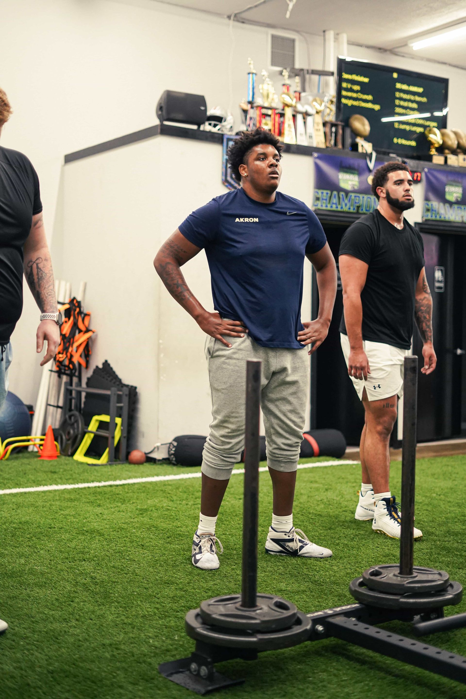 Three athletes in a gym stand near sleds on turf. One man in blue looks up, hands on hips.
