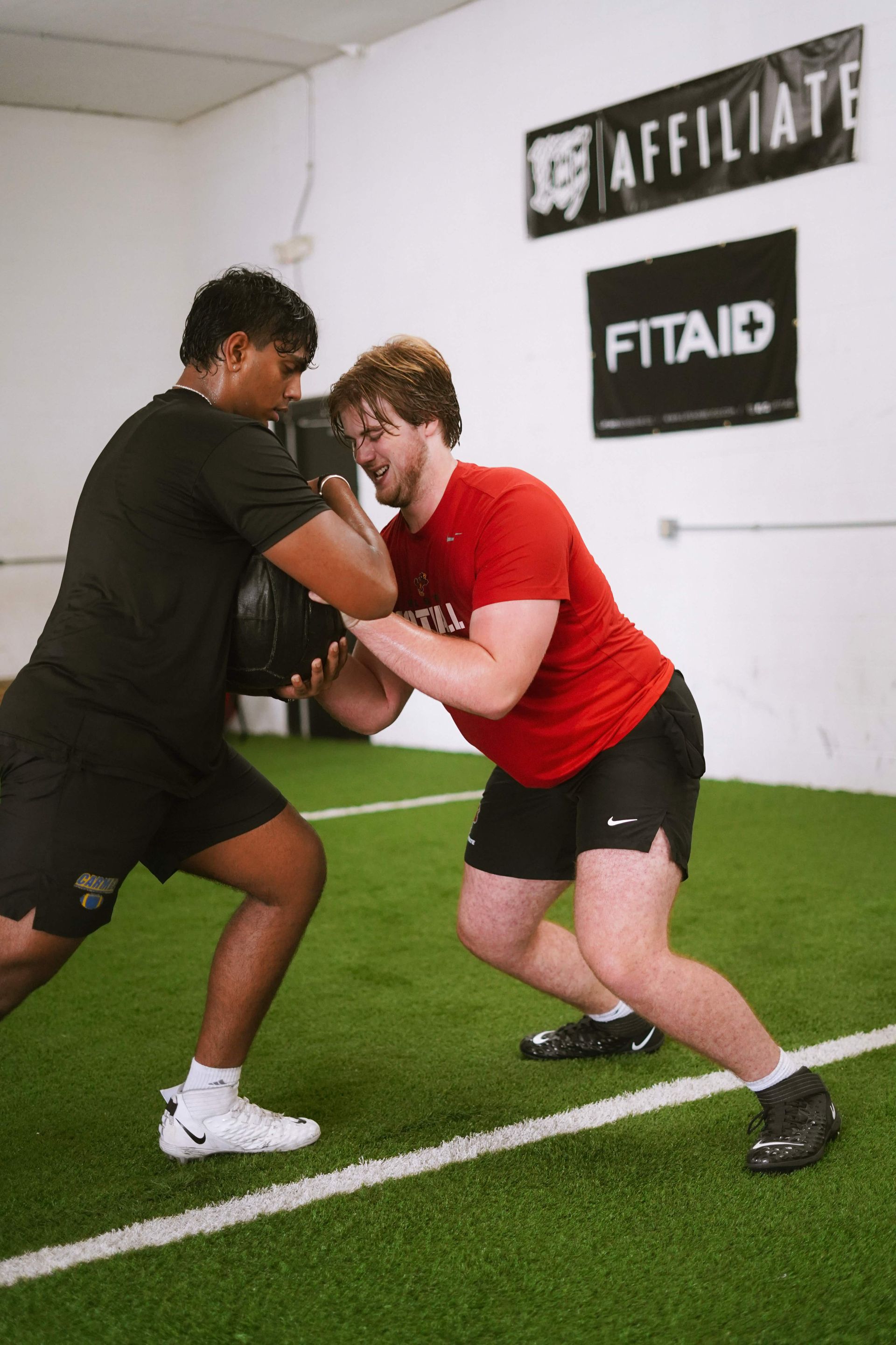 Two men in a gym train with a medicine ball. One in black, the other in red. They face each other on green turf.